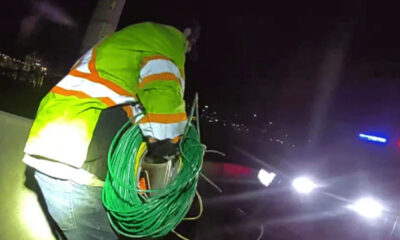 Utility worker in a high-visibility jacket coils a green cable beside a car with flashing red and blue emergency lights at night.