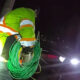 Utility worker in a high-visibility jacket coils a green cable beside a car with flashing red and blue emergency lights at night.