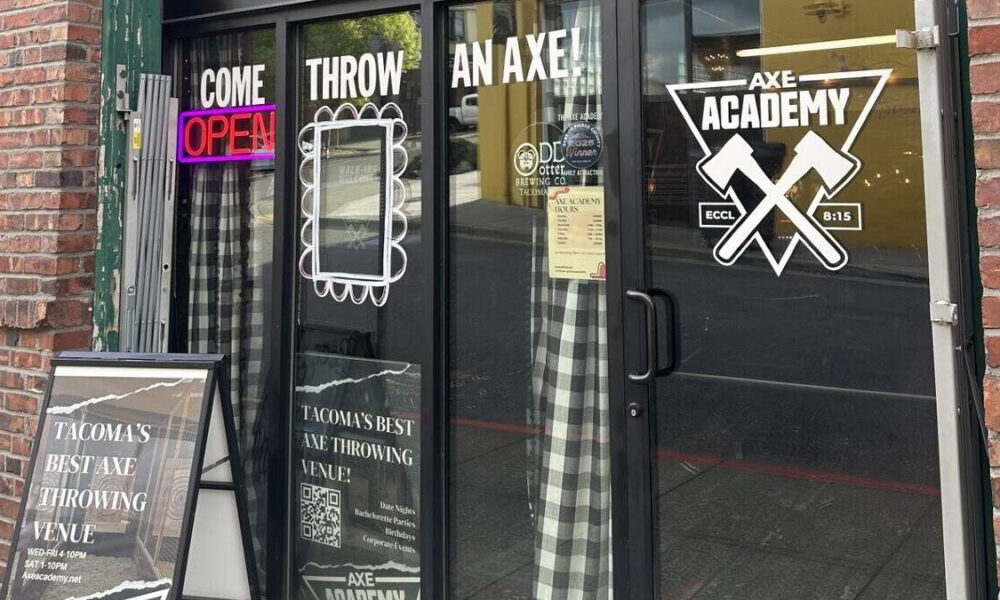 Storefront advertising an axe-throwing venue: glass doors say 'Come Throw An Axe' with a neon 'OPEN' sign and an 'AXE ACADEMY' logo; a sidewalk sign outside sits on brick.