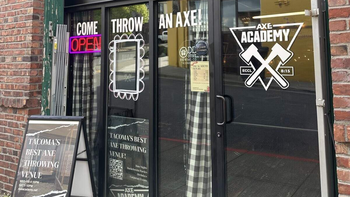 Storefront advertising an axe-throwing venue: glass doors say 'Come Throw An Axe' with a neon 'OPEN' sign and an 'AXE ACADEMY' logo; a sidewalk sign outside sits on brick.