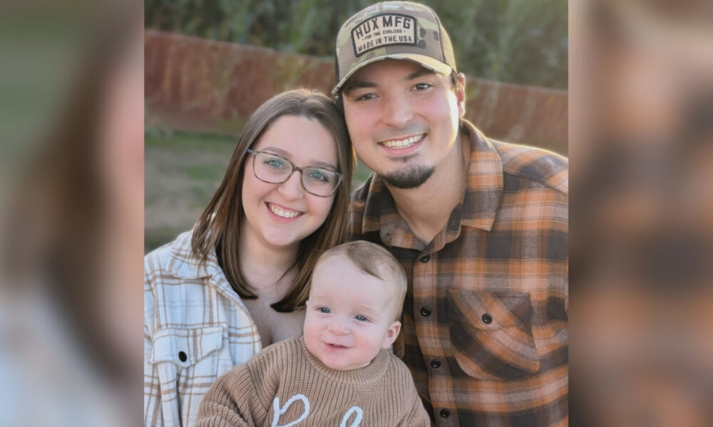 Smiling couple with their baby outdoors, posing closely together in casual flannel shirts and a cap behind them