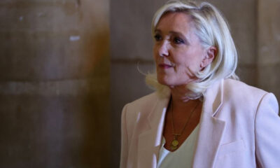 Senior woman with short blonde hair wearing a light blazer and gold pendant necklace, looking to the right.