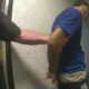 Police officer escorts a man in a blue shirt down a narrow hallway, with the detainee's hands restrained behind his back.