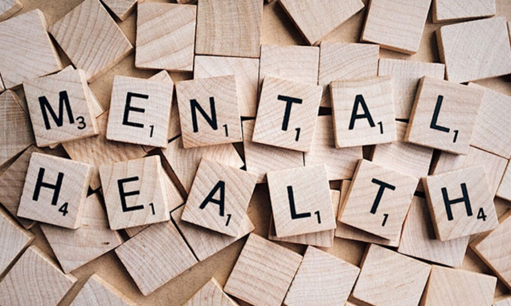 Wooden Scrabble tiles spell 'MENTAL HEALTH' on a pale wooden surface.