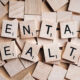 Wooden Scrabble tiles spell 'MENTAL HEALTH' on a pale wooden surface.