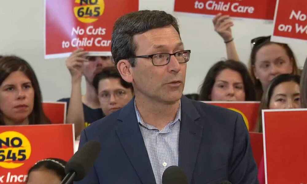 Man in a navy blazer and glasses speaks at a press conference with red protest signs behind him reading 'No Cuts to Child Care'.