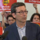 Man in a navy blazer and glasses speaks at a press conference with red protest signs behind him reading 'No Cuts to Child Care'.