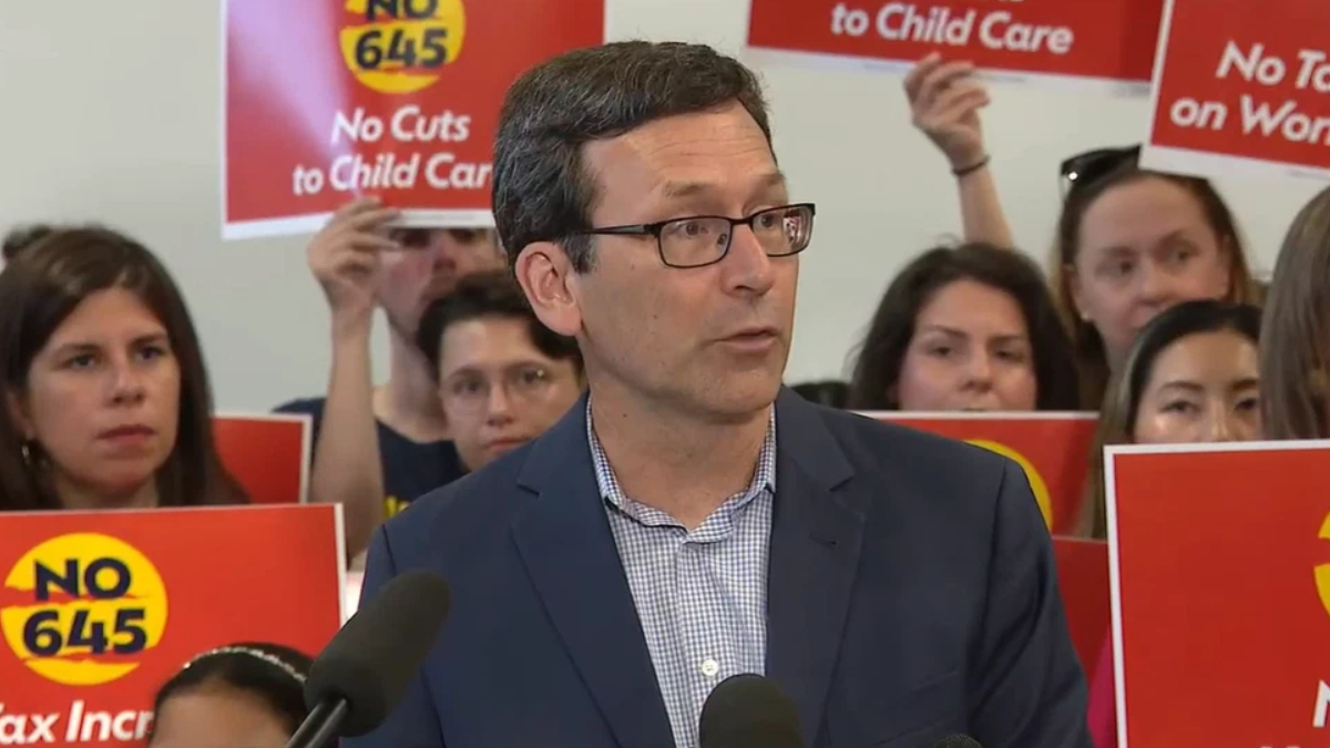 Man in a navy blazer and glasses speaks at a press conference with red protest signs behind him reading 'No Cuts to Child Care'.