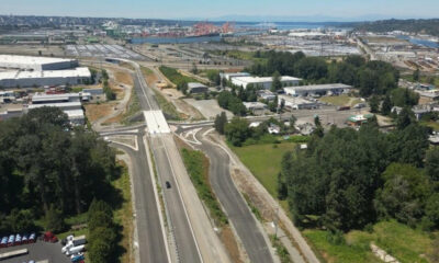 Aerial view of a multi‑lane highway interchange under construction near an industrial area with a port in the distance.