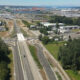 Aerial view of a multi‑lane highway interchange under construction near an industrial area with a port in the distance.