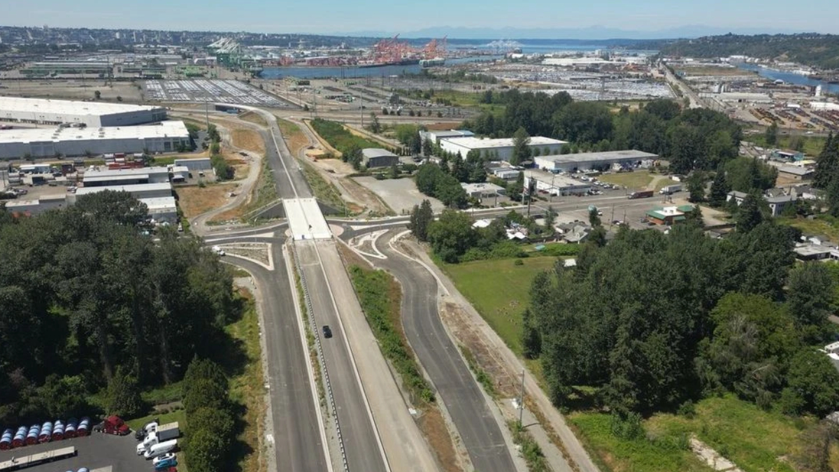 Aerial view of a multi‑lane highway interchange under construction near an industrial area with a port in the distance.