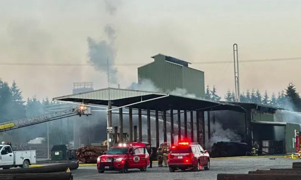 Industrial building on fire with smoke rising; firefighters and red emergency vehicles on scene.