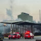Industrial building on fire with smoke rising; firefighters and red emergency vehicles on scene.