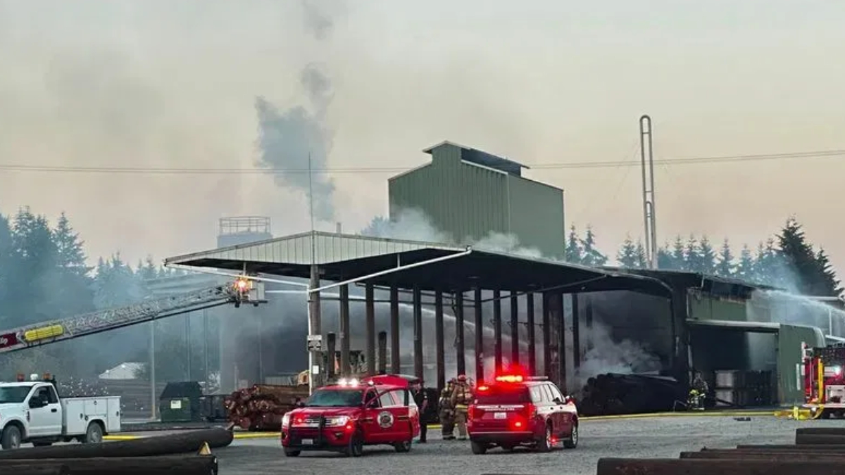 Industrial building on fire with smoke rising; firefighters and red emergency vehicles on scene.