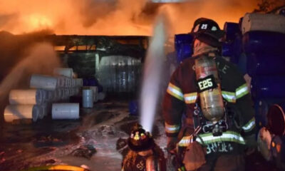 Firefighter in turnout gear sprays water at a blazing storage area with blue drums and white pipes nearby, at night.2024-07-22T00:00:00Z