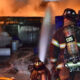 Firefighter in turnout gear sprays water at a blazing storage area with blue drums and white pipes nearby, at night.2024-07-22T00:00:00Z