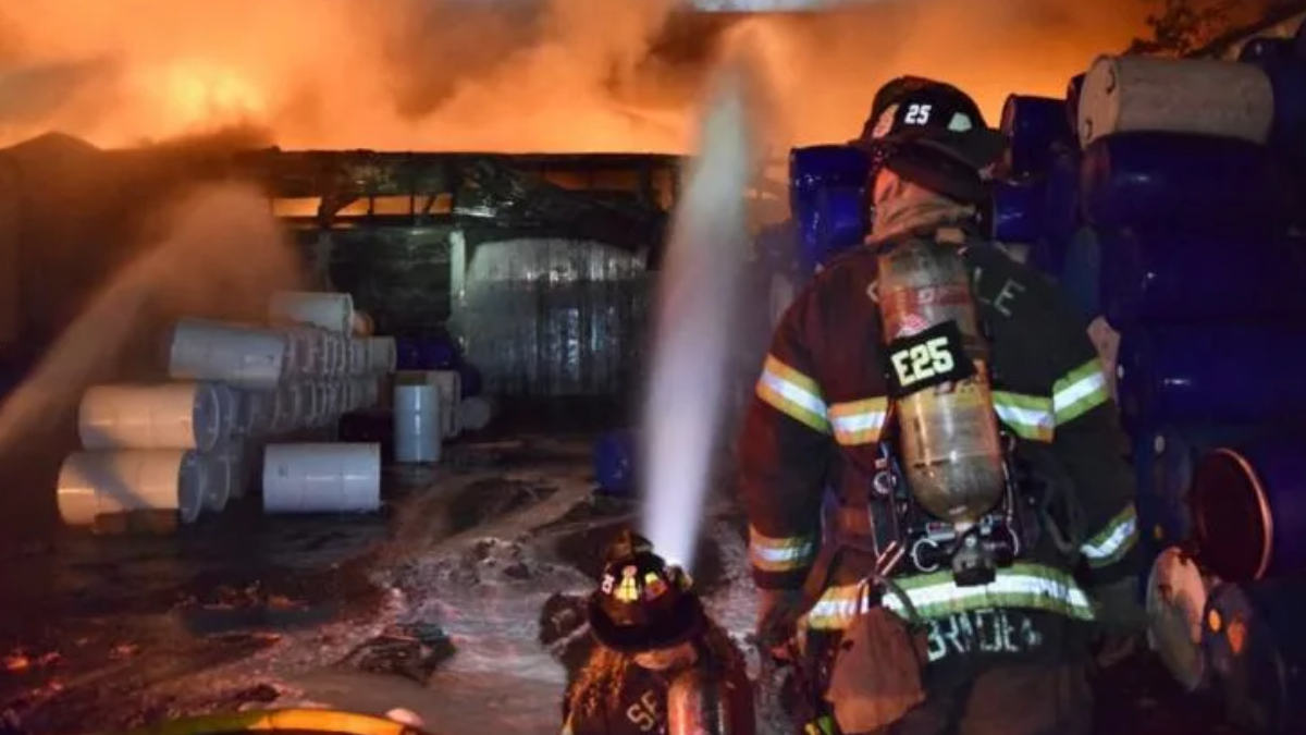 Firefighter in turnout gear sprays water at a blazing storage area with blue drums and white pipes nearby, at night.2024-07-22T00:00:00Z
