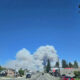 Suburban street with houses and cars as a huge white smoke plume rises from distant hills under a clear blue sky.