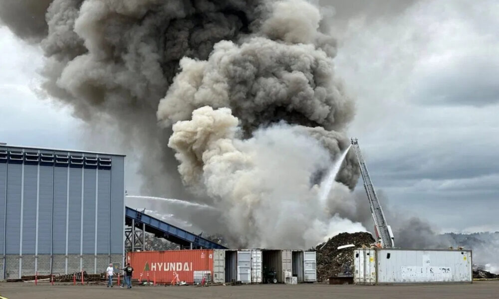 Industrial site fire with thick black and gray smoke billowing from a pile of material; a firefighter ladder truck sprays water onto the flames.