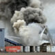 Industrial site fire with thick black and gray smoke billowing from a pile of material; a firefighter ladder truck sprays water onto the flames.