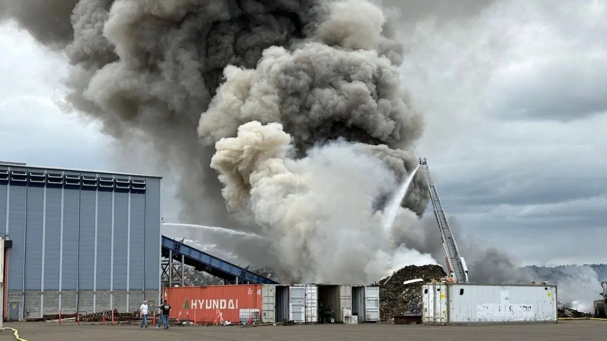 Industrial site fire with thick black and gray smoke billowing from a pile of material; a firefighter ladder truck sprays water onto the flames.