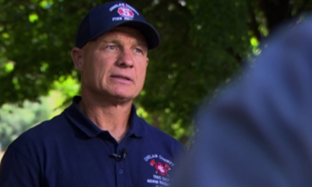 Man in a dark navy polo and cap with a red emblem speaks outdoors, microphone clipped to his shirt, blurred greenery in the background.
