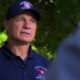 Man in a dark navy polo and cap with a red emblem speaks outdoors, microphone clipped to his shirt, blurred greenery in the background.