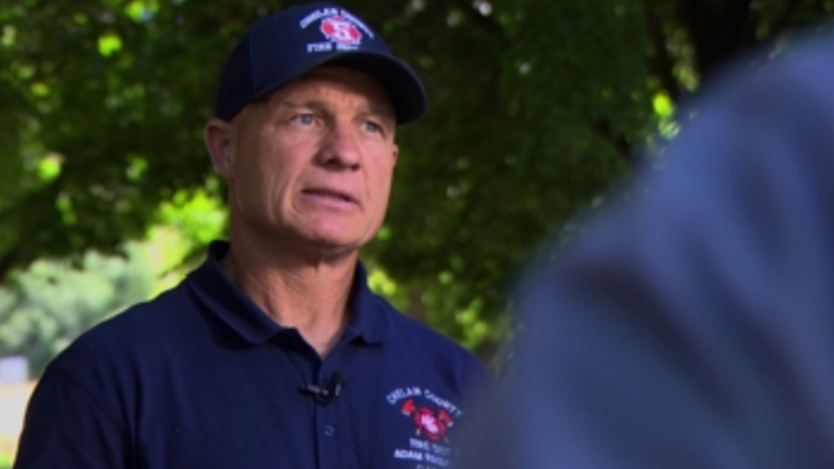 Man in a dark navy polo and cap with a red emblem speaks outdoors, microphone clipped to his shirt, blurred greenery in the background.