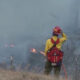 Firefighter in yellow shirt and red helmet stands amid smoke and flames on a grassy hillside, holding a hose near a fence.