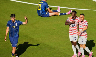 Soccer scene on a green field: a blue-team player (number 19) walks left while two players in red-and-white stripes (numbers 20 and 2) celebrate together; another blue-team player (number 21) sits on the ground in the background.