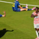Soccer scene on a green field: a blue-team player (number 19) walks left while two players in red-and-white stripes (numbers 20 and 2) celebrate together; another blue-team player (number 21) sits on the ground in the background.