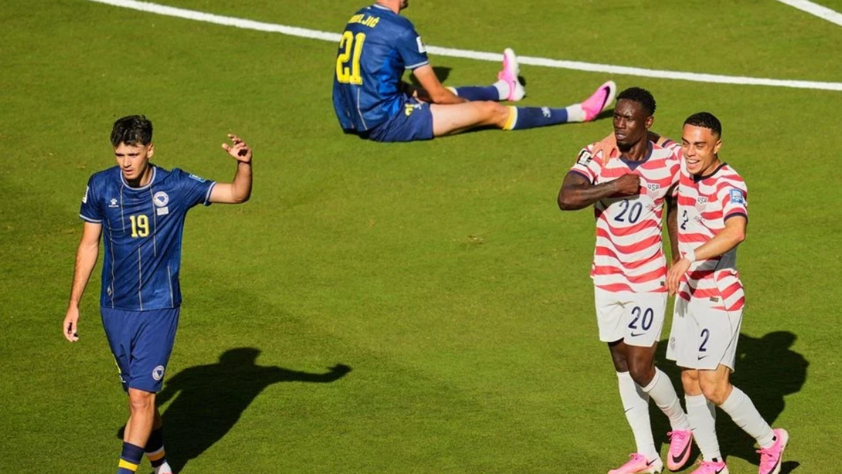 Soccer scene on a green field: a blue-team player (number 19) walks left while two players in red-and-white stripes (numbers 20 and 2) celebrate together; another blue-team player (number 21) sits on the ground in the background.