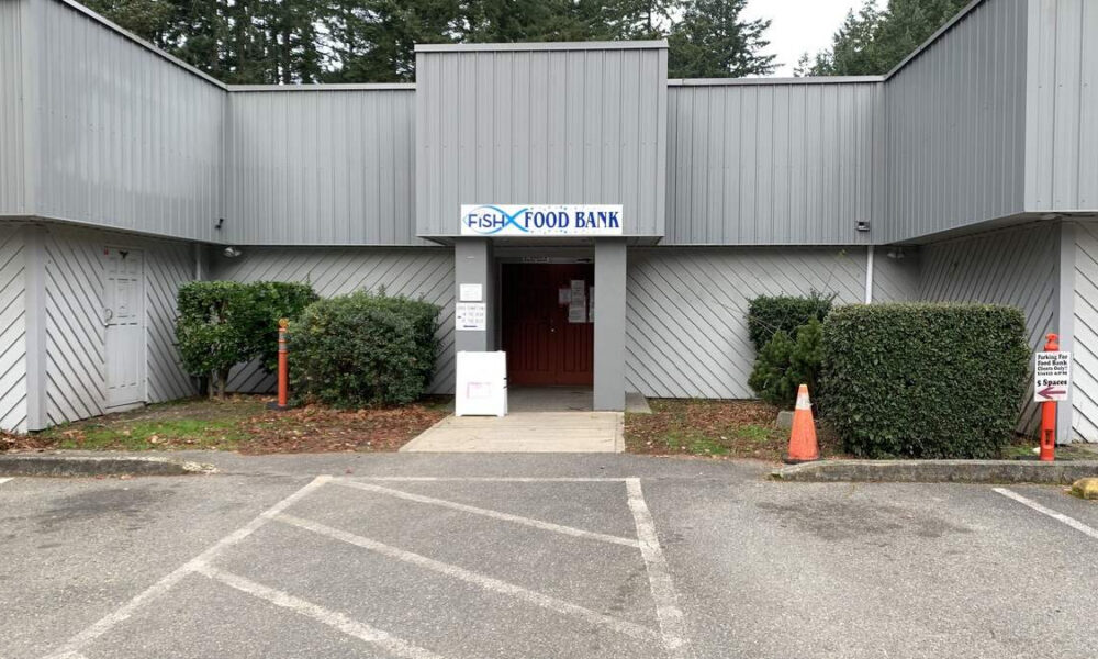 Front entrance of a gray metal building labeled 'Fish Food Bank,' with a red door and a concrete walkway, framed by hedges and a parking lot in front.
