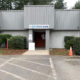 Front entrance of a gray metal building labeled 'Fish Food Bank,' with a red door and a concrete walkway, framed by hedges and a parking lot in front.