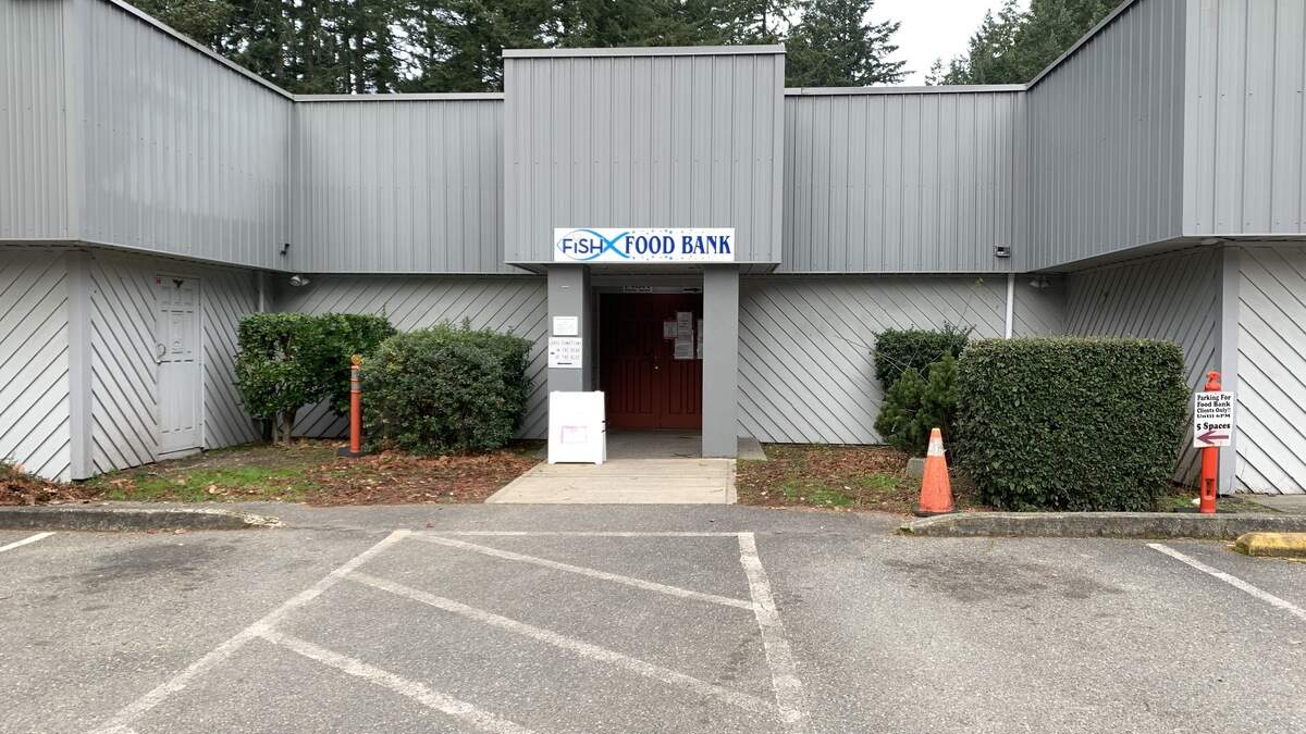 Front entrance of a gray metal building labeled 'Fish Food Bank,' with a red door and a concrete walkway, framed by hedges and a parking lot in front.