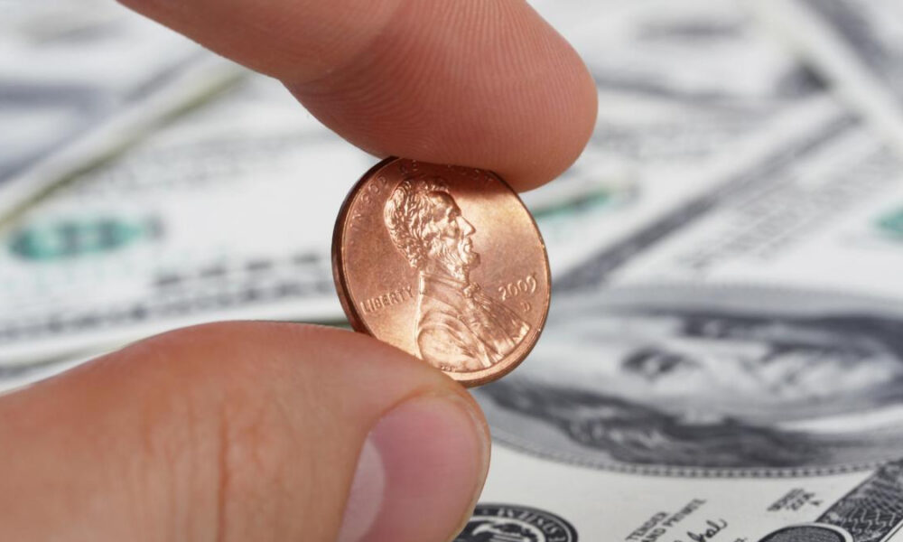Close-up of a penny held between a thumb and forefinger, with US dollar bills blurred in the background.
