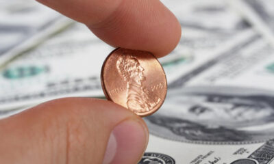 Close-up of a penny held between a thumb and forefinger, with US dollar bills blurred in the background.