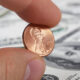Close-up of a penny held between a thumb and forefinger, with US dollar bills blurred in the background.