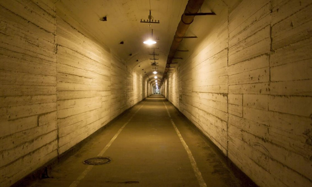 Long concrete tunnel with arched ceiling, yellowish lighting, and a central walkway fading to a distant light.