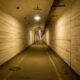 Long concrete tunnel with arched ceiling, yellowish lighting, and a central walkway fading to a distant light.