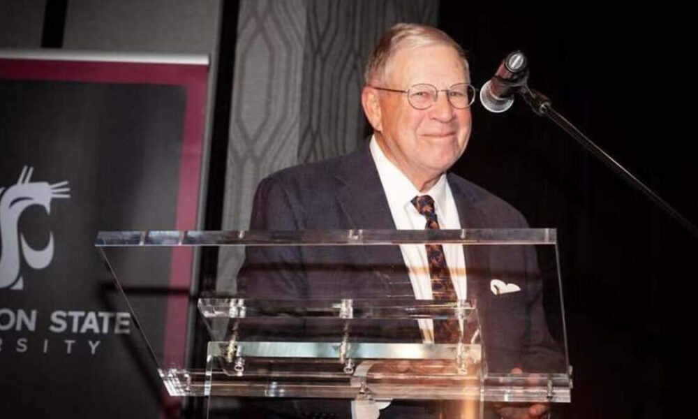 Older man in a suit at a clear lectern giving a speech, microphone overhead, with a banner in the background.