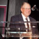 Older man in a suit at a clear lectern giving a speech, microphone overhead, with a banner in the background.