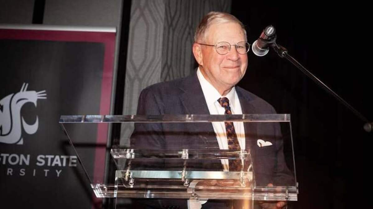 Older man in a suit at a clear lectern giving a speech, microphone overhead, with a banner in the background.