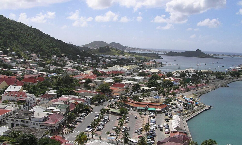 Aerial view of a coastal town built on hills, with colorful red-roofed buildings, a busy waterfront, and a blue bay.