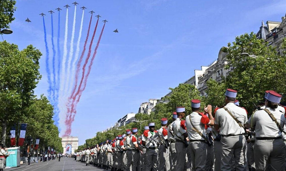Planes fly in a line releasing blue, white, and red smoke over a tree-lined parade route as uniformed soldiers stand in formation along the street.