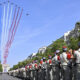 Planes fly in a line releasing blue, white, and red smoke over a tree-lined parade route as uniformed soldiers stand in formation along the street.