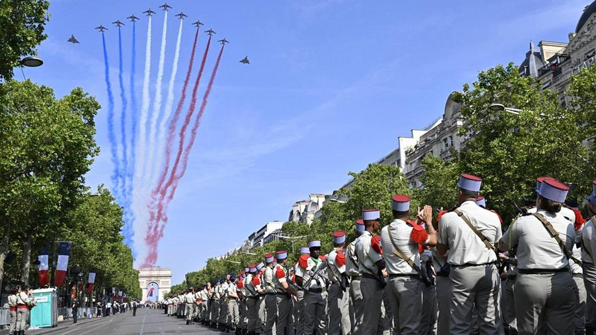 Planes fly in a line releasing blue, white, and red smoke over a tree-lined parade route as uniformed soldiers stand in formation along the street.