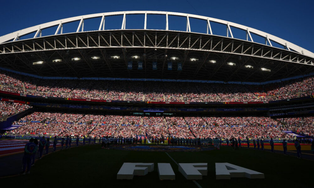 Arena filled with tens of thousands of spectators during a FIFA event, view of the stadium stands under a bright blue sky, large 'FIFA' lettering on the field centerpiece.
