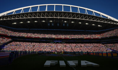 Arena filled with tens of thousands of spectators during a FIFA event, view of the stadium stands under a bright blue sky, large 'FIFA' lettering on the field centerpiece.