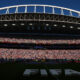 Arena filled with tens of thousands of spectators during a FIFA event, view of the stadium stands under a bright blue sky, large 'FIFA' lettering on the field centerpiece.